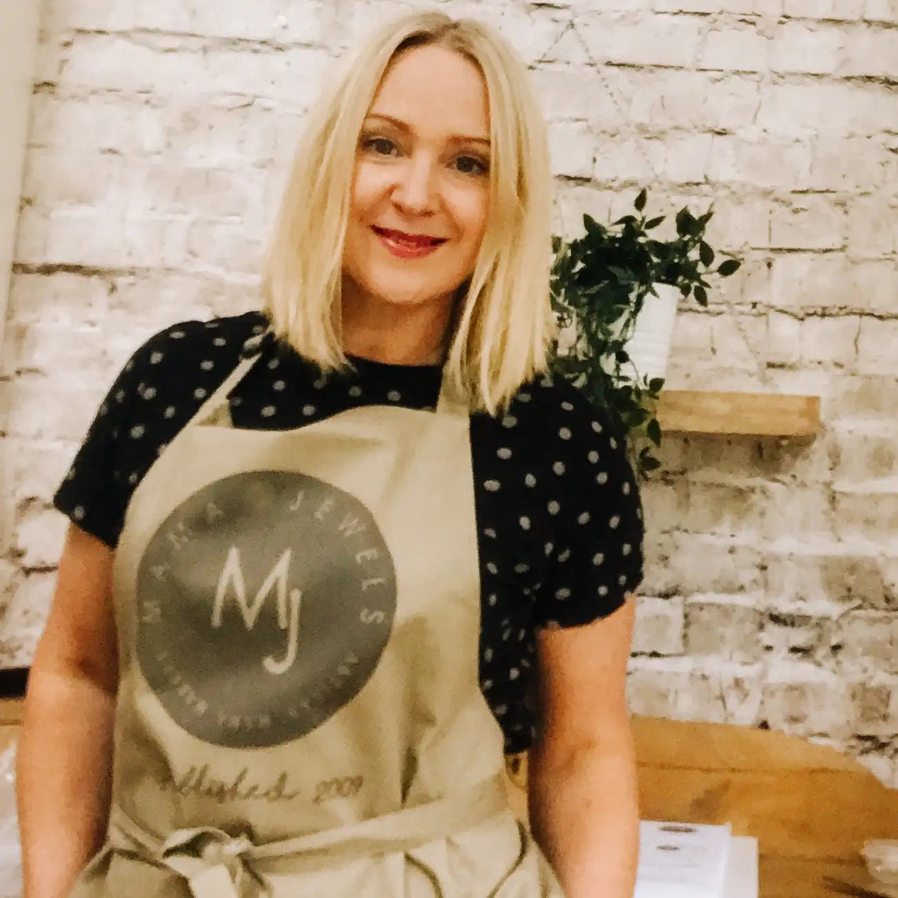 Beige apron with a circular dark gray logo featuring ’mj’ and the text ’mack jewels’ around it, tied at the waist.