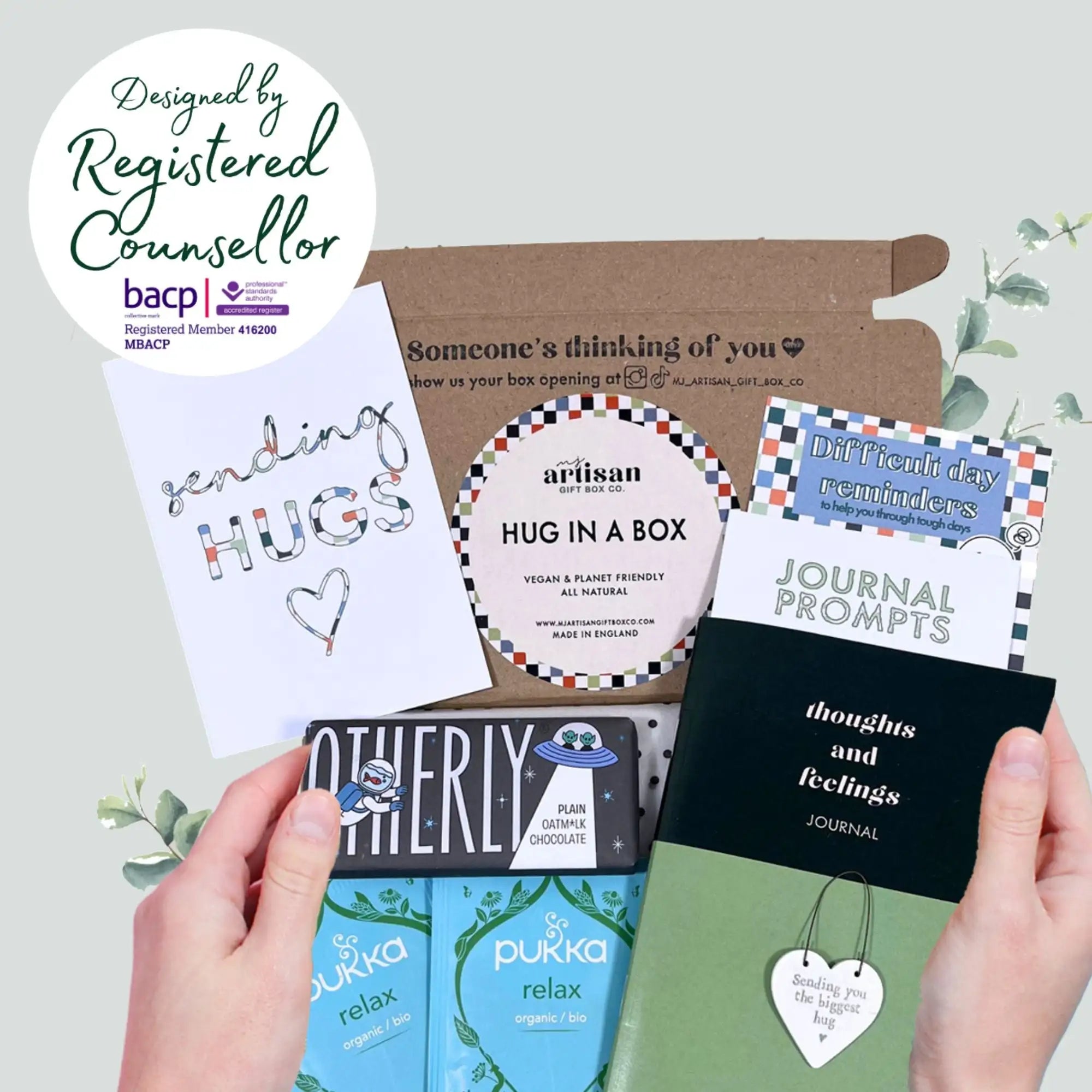 A black journal with white text on the cover, titled ’thoughts and feelings,’ held in hands alongside other items from a self-care gift box.