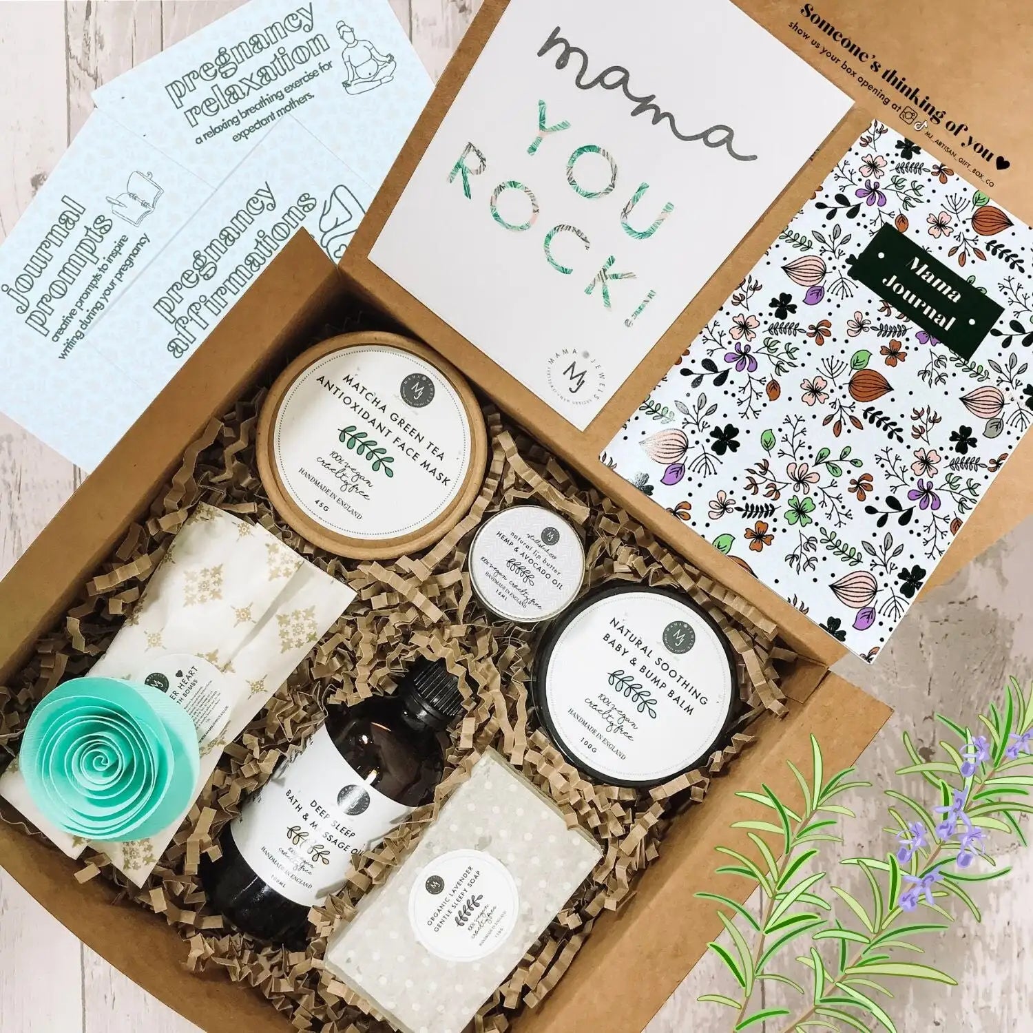 A brown cardboard gift box filled with pregnancy care items including a matcha green tea face mask, baby & bump balm, bath oil, and a floral-patterned journal.